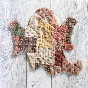 May include: A ghost-shaped quilted fabric ornament constructed from various patchwork squares featuring floral and geometric patterns in muted tones of brown, tan, red, green, and yellow, with two eye cutouts and one mouth cutout.