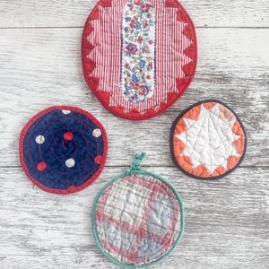 May include: A collection of four quilted pot holders in various shapes and patterns. One is oval with red and white stripes and floral center. Another is round with blue fabric and white polka dots. The third is round with orange and white starburst design. The last is round with a plaid pattern.