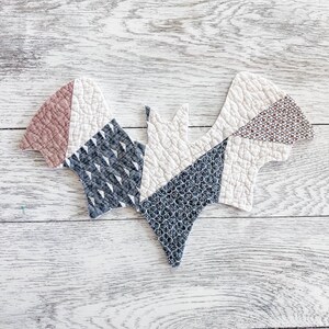 May include: A quilted fabric bat ornament featuring a patchwork design. The bat is constructed from various fabric pieces in shades of cream, dusty rose, and dark charcoal gray, each displaying distinct geometric and floral patterns with visible stitching.