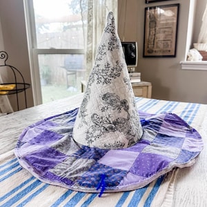May include: A conical hat featuring a white fabric crown with a black floral print and a wide, circular brim constructed from a patchwork of purple, lavender, and blue fabric squares. The entire piece is quilted with visible stitching throughout.