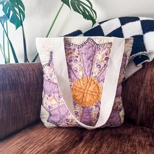 K&ouml;nnte beinhalten: Eine Tragetasche mit floralem Muster in Lila und Gelb mit wei&szlig;en Henkeln. Die Tasche ist aus gestepptem Stoff und hat ein Sternburst-Design.
