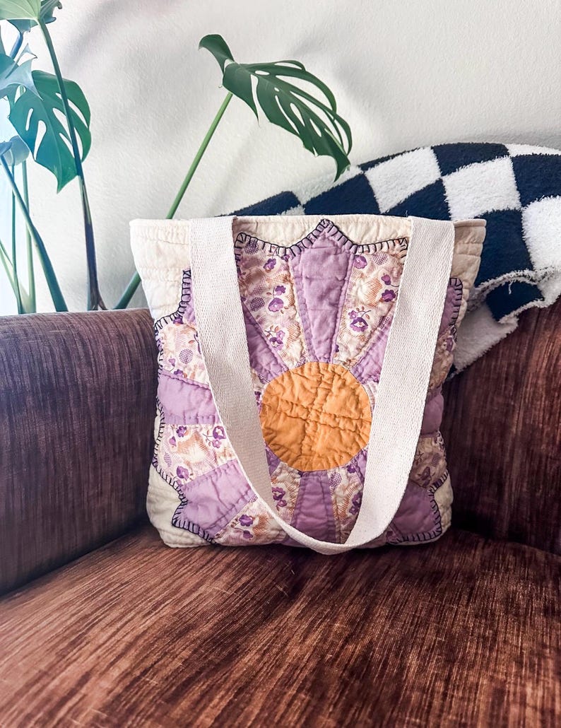 K&ouml;nnte beinhalten: Eine Tragetasche mit floralem Muster in Lila und Gelb mit wei&szlig;en Henkeln. Die Tasche ist aus gestepptem Stoff und hat ein Sternburst-Design.