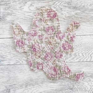May include: A ghost-shaped fabric cutout featuring a quilted texture with a floral pattern in shades of pink, green, and beige. The ghost has two oval-shaped eye cutouts and a small circular mouth cutout, with stitched edges defining its form.