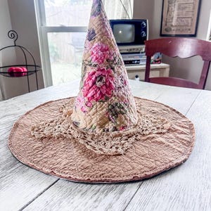 May include: A conical hat featuring a quilted tan fabric with a pink and green floral pattern on the crown. The wide, flat brim is made of solid tan quilted fabric, accented by a decorative beige crocheted lace trim around the base of the cone.
