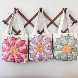 K&ouml;nnte beinhalten: Drei gesteppte Tragetaschen mit Blumendesigns. Die Taschen h&auml;ngen an einem Holzgestell. Die Blumen sind rosa, lila und gr&uuml;n.