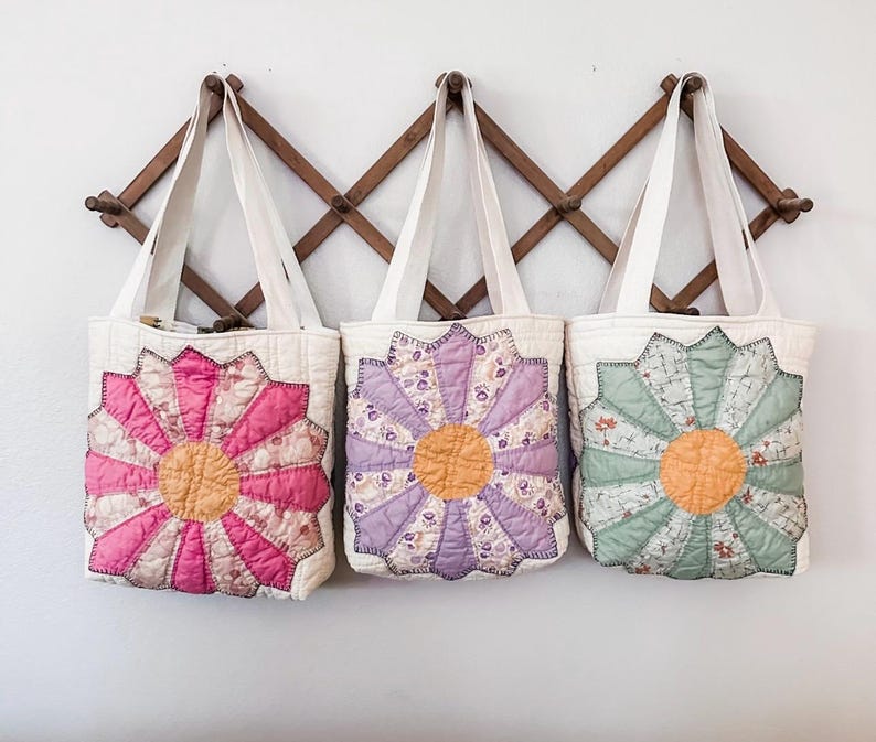 K&ouml;nnte beinhalten: Drei gesteppte Tragetaschen mit Blumendesigns. Die Taschen h&auml;ngen an einem Holzgestell. Die Blumen sind rosa, lila und gr&uuml;n.