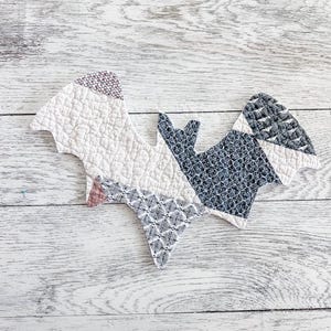 May include: A quilted fabric bat ornament featuring a patchwork design. The left wing is cream with a textured pattern, while the right wing and body consist of dark grey fabric with white floral and geometric prints, finished with stitched edges.