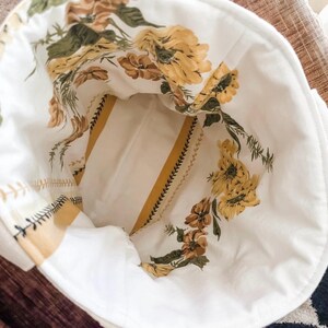 K&ouml;nnte beinhalten: Eine wei&szlig;e Stofftasche mit gelbem und gr&uuml;nem Blumendruck. Die Tasche hat ein gestreiftes Futter und einen Rei&szlig;verschluss.