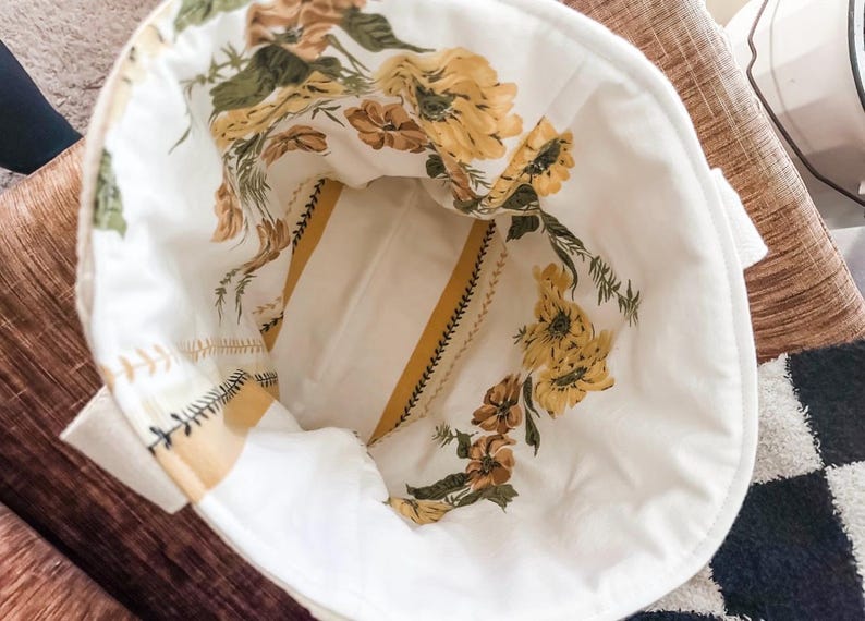 K&ouml;nnte beinhalten: Eine wei&szlig;e Stofftasche mit gelbem und gr&uuml;nem Blumendruck. Die Tasche hat ein gestreiftes Futter und einen Rei&szlig;verschluss.