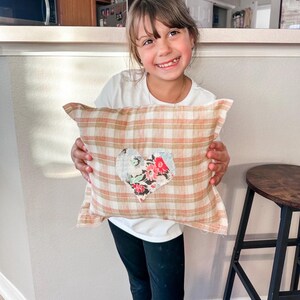 May include: A plaid pillow with a heart-shaped floral patch. The pillow features a cream, peach, and brown plaid pattern. The heart patch is made of floral fabric with red, pink, and blue flowers. The pillow is rectangular.