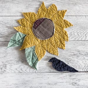 May include: A quilted fabric sunflower with mustard yellow petals, a dark brown plaid center, and two sage green leaves. Beside the flower is a small, dark navy blue quilted fabric bird shape. Both items feature visible stitched textures.