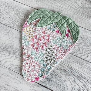 May include: A strawberry-shaped quilted fabric mat featuring a floral pattern in shades of pink, white, and light blue. The top section consists of a sage green quilted leaf element with a stitched grid texture, contrasting with the floral body.