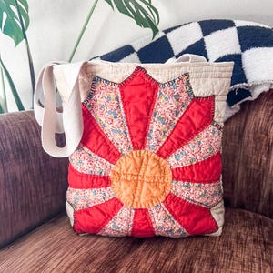 K&ouml;nnte beinhalten: Eine rote, wei&szlig;e und orangefarbene gesteppte Tragetasche mit Blumenmuster. Die Tasche hat ein gro&szlig;es, kreisf&ouml;rmiges Design in der Mitte, mit einem kleineren Kreis in der Mitte. Die Tasche hat zwei lange Tragegurte.