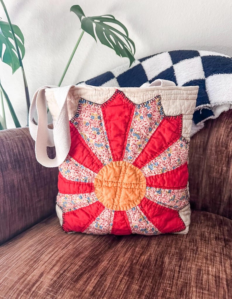 K&ouml;nnte beinhalten: Eine rote, wei&szlig;e und orangefarbene gesteppte Tragetasche mit Blumenmuster. Die Tasche hat ein gro&szlig;es, kreisf&ouml;rmiges Design in der Mitte, mit einem kleineren Kreis in der Mitte. Die Tasche hat zwei lange Tragegurte.