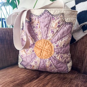 K&ouml;nnte beinhalten: Eine violette und gelbe Patchwork-Tragetasche mit Blumendesign und einem Canvas-Trageriemen. Die Tasche ist aus verschiedenen Stoffen gefertigt und hat ein gestepptes Design.