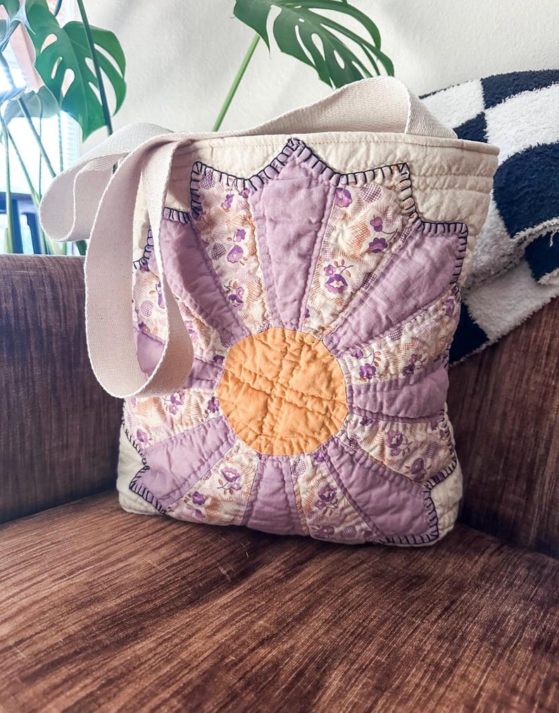 K&ouml;nnte beinhalten: Eine violette und gelbe Patchwork-Tragetasche mit Blumendesign und einem Canvas-Trageriemen. Die Tasche ist aus verschiedenen Stoffen gefertigt und hat ein gestepptes Design.