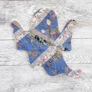 May include: A ghost-shaped fabric ornament constructed from quilted patchwork pieces. The design features a blue floral print fabric combined with strips of beige and brown plaid fabric. It has two circular cutouts for eyes and one for a mouth.
