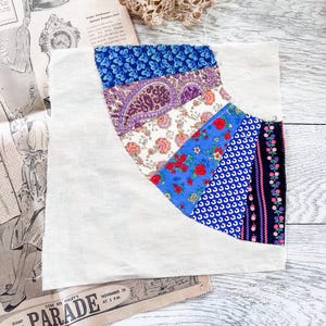 May include: A square fabric quilt block featuring a curved fan design made of multiple patterned strips. The fabric includes blue floral, purple paisley, cream floral, red floral, blue geometric, and black floral prints on a plain white background.
