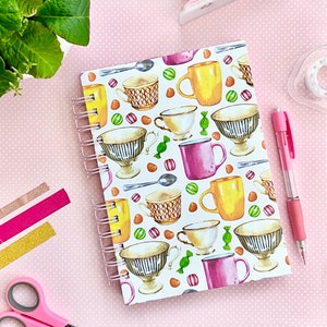 Pode incluir: Um caderno branco de espiral com um padrão colorido de xícaras de chá e doces. O caderno tem um fundo branco com um padrão repetitivo de xícaras de chá, pires e doces.
