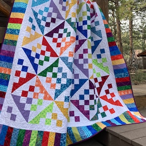 May include: A colorful quilt with a white background and a variety of geometric patterns in shades of purple, blue, green, orange, and red. The quilt is bordered with a rainbow stripe pattern.