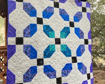 May include: A purple, white, and blue quilt with a geometric pattern. The quilt is hanging on a wooden structure.