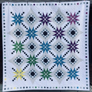 May include: A white quilt with a geometric pattern of stars and circles in various colors, including green, blue, purple, yellow, and black.