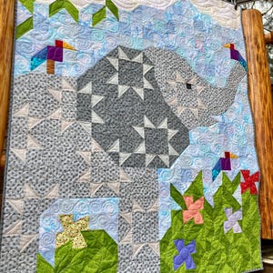 Puede incluir: Una colcha gris y blanca con un patrón geométrico. La colcha presenta un gran elefante gris con un contorno blanco y un fondo azul y blanco. La colcha está rodeada de hojas verdes y flores en tonos de rosa, morado y rojo.