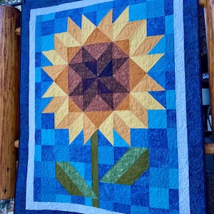 Op de afbeelding: Een gewatteerde wandhanger met een zonnebloemontwerp. De zonnebloem heeft een bruin hart, gele en oranje bloemblaadjes en groene bladeren en stengel. De achtergrond is een blauw patchworkpatroon met een witte rand.