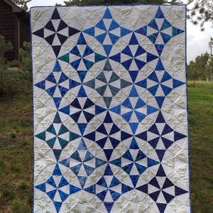 May include: A white quilt with a blue and white geometric pattern. The quilt is made up of many small triangles that form a larger star shape. The quilt is hanging on a clothesline in a grassy area.