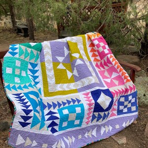 May include: A colorful quilt with a patchwork design featuring geometric shapes in shades of blue, green, yellow, pink, and purple. The quilt is made with a variety of fabrics and textures, and it is displayed on a wooden bench.