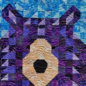 Pode incluir: Uma colcha de retalhos roxa e azul com um design de rosto de urso. O nariz e a boca do urso são de cor castanho claro.