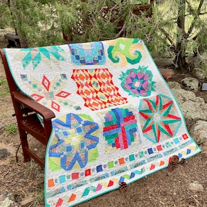 Op de afbeelding: Een handgemaakte quilt met een witte achtergrond en kleurrijke geometrische patronen. De quilt heeft verschillende vormen en ontwerpen in tinten blauw, groen, rood en geel. Het is buiten over een houten stoel gedrapeerd.