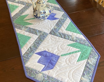 May include: A quilted table runner with a geometric pattern in shades of green, purple, and white. The runner has a scalloped edge and is decorated with a floral print. A blue and white teapot sits on top of the runner.