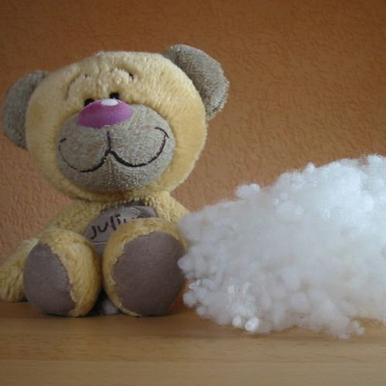 May include: A plush teddy bear with a pink nose and a smile, sitting next to a pile of white stuffing. The bear is light brown and has the name "Julia" written on its chest.