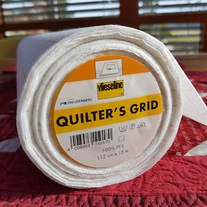 Könnte beinhalten: Eine Rolle weißer Quilting-Gitterstoff mit einem gelben Etikett, auf dem "vlieseline QUILTER'S GRID" steht, sowie Pflegehinweise und Produktinformationen. Das Etikett enthält auch den Text "FREUDENBERG" und einen Barcode.