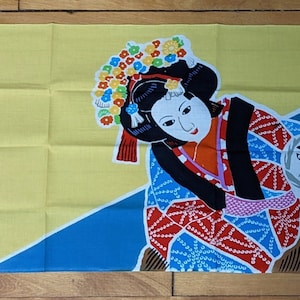 May include: A rectangular, yellow, blue, and red Japanese textile featuring a Kabuki theater scene. The design includes two figures in traditional attire, with one wearing a colorful headdress. The textile has a small, square stamp in the lower left corner.
