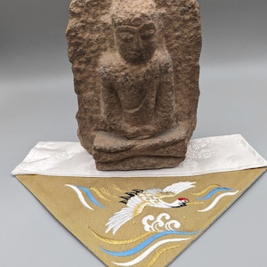 Puede incluir: Una escultura de piedra de un Buda sentado sobre un cojín bordado en blanco y dorado con un diseño de grúa. El cojín tiene un fondo blanco con bordados dorados y azules.
