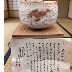 May include: A handcrafted ceramic tea bowl with a textured, cratered surface featuring a white glaze and reddish-brown accents. The bowl rests on a wooden box with a lid, accompanied by a piece of paper with Japanese calligraphy and a red seal.