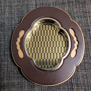 Könnte beinhalten: Ein dekorativer Untersetzer mit einem braunen, gewellten Rand und einem gold-beigen Akzent. Die Mitte zeigt ein geometrisches Muster in Gold und Schwarz. Der Untersetzer hat einen silbernen Rand und einen beigen Rand.