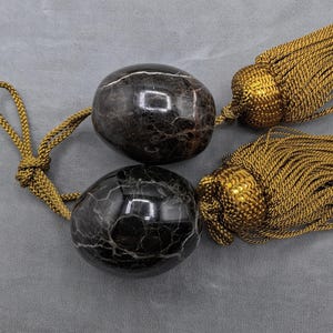 May include: Two decorative black marble egg-shaped tassels with gold-colored rope and tassels. The marble has white veining. The tassels are likely used for curtains or home decor.