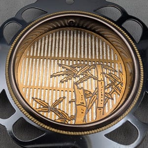 Pode incluir: Uma base decorativa com um anel exterior prateado e um centro dourado. O centro apresenta um desenho de bambu com linhas verticais e caules e folhas de bambu. A base tem um design detalhado e vazado.