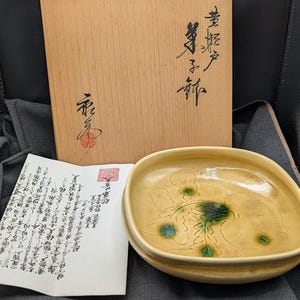 May include: A square ceramic dish with a light yellow glaze and green floral design. The dish is in a wooden box with Japanese characters on the lid.
