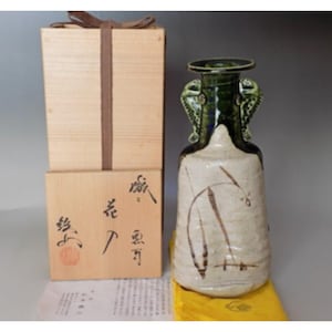 May include: A ceramic vase with a green glaze and a white body. The vase has two handles shaped like stylised fish. The vase is decorated with a brown and white design of reeds. The vase is sitting on a yellow cloth and is in a wooden box with Japanese characters.