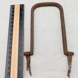 以下が含まれることがあります： U字型のフレームと下部に2本のツメが付いた、さび付いた金属製の工具。この工具は約15cmの長さで、定規で測定されています。
