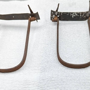 Op de afbeelding: Twee roestige U-vormige metalen beugels met een plat metalen plaatje bovenaan. De beugels zijn met twee klinknagels aan elke kant aan de plaat bevestigd.