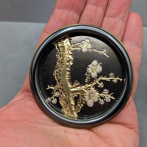 May include: A black and gold decorative coaster with a floral design. The coaster features a gold tree branch with white flowers against a black background, framed by a gold and black border.