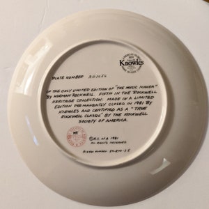 May include: A white ceramic plate with a brown border and a printed inscription. The inscription reads "PLATE NUMBER AG1686 OF THE ONLY LIMITED EDITION OF "THE MUSIC MAKER" BY NORMAN ROCKWELL. FIFTH IN THE ROCKWELL HERITAGE COLLECTION. MADE IN A LIMITED EDITION PERMANENTLY CLOSED IN 1981 BY KNOWLES AND CERTIFIED AS A "TRUE ROCKWELL CLASSIC" BY THE ROCKWELL SOCIETY OF AMERICA. R.S. of A 1981 ALL RIGHTS RESERVED Bradex Number: 84-R70-3-5"