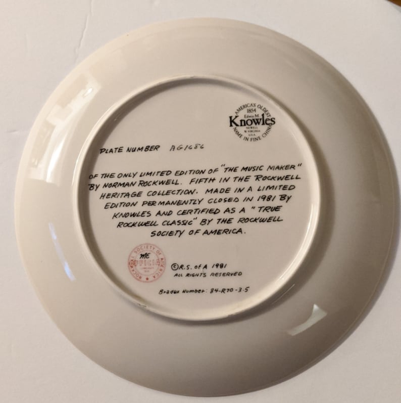 May include: A white ceramic plate with a brown border and a printed inscription. The inscription reads "PLATE NUMBER AG1686 OF THE ONLY LIMITED EDITION OF "THE MUSIC MAKER" BY NORMAN ROCKWELL. FIFTH IN THE ROCKWELL HERITAGE COLLECTION. MADE IN A LIMITED EDITION PERMANENTLY CLOSED IN 1981 BY KNOWLES AND CERTIFIED AS A "TRUE ROCKWELL CLASSIC" BY THE ROCKWELL SOCIETY OF AMERICA. R.S. of A 1981 ALL RIGHTS RESERVED Bradex Number: 84-R70-3-5"