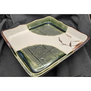 Puede incluir: Una bandeja de servir cuadrada de cerámica con fondo blanco y diseños verdes y marrones. La bandeja tiene una superficie texturizada y un borde elevado.