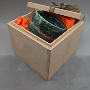 May include: A handcrafted ceramic bowl with a speckled green and brown glaze, nestled inside a light-colored wooden box. The box is lined with orange fabric and topped with a lid tied with a green and brown ribbon. The bowl is a unique piece of art.